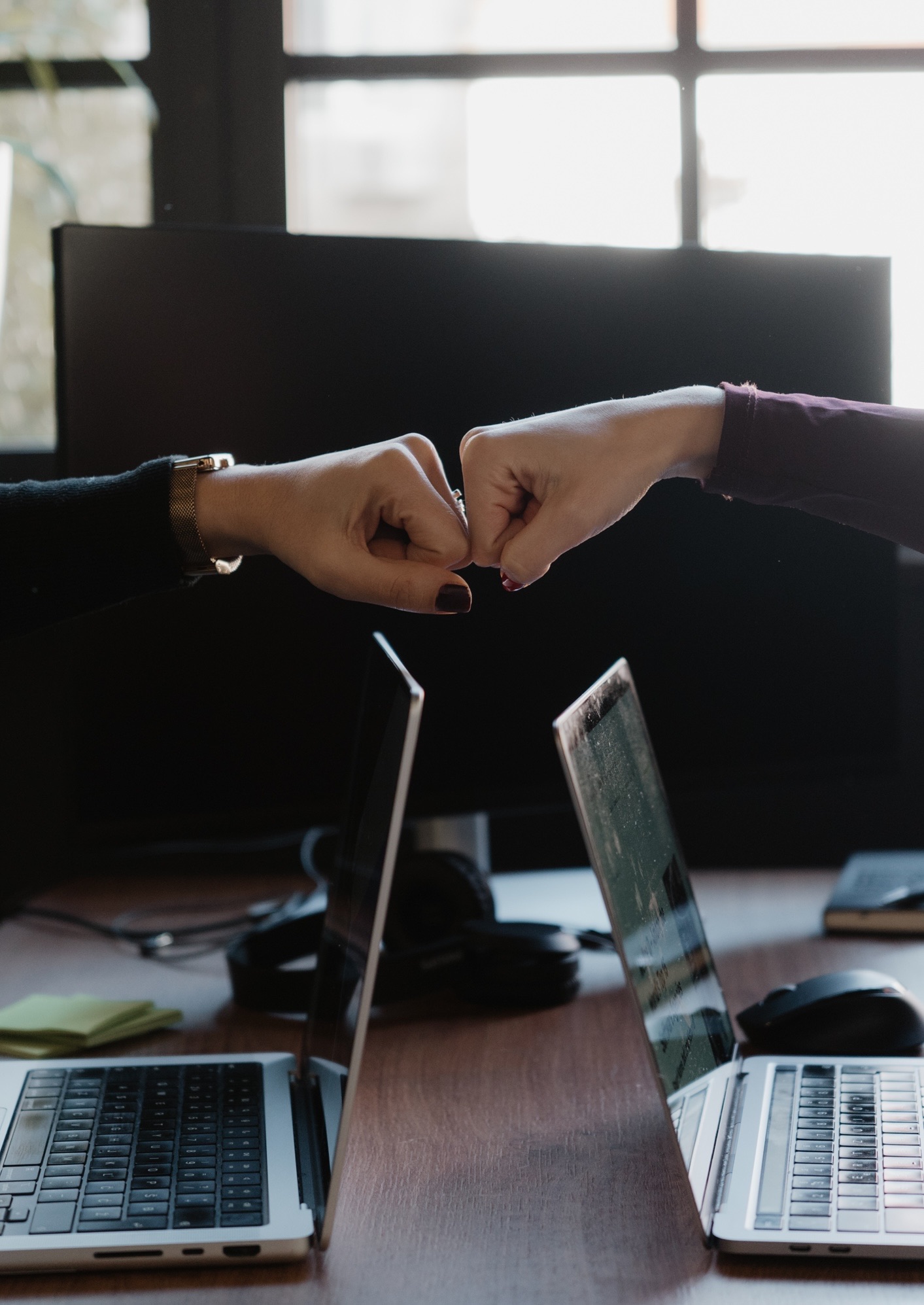 Colleagues fist-bumping across laptops in a modern workspace, symbolising teamwork and cooperation.